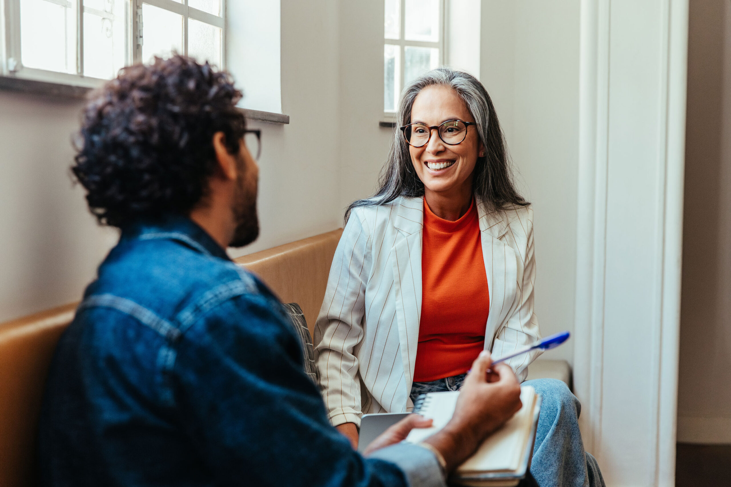 FounderSmall A smiling woman with glasses and gray hair in an orange top and pinstripe blazer talks to a man in a denim jacket who is writing in a notebook.