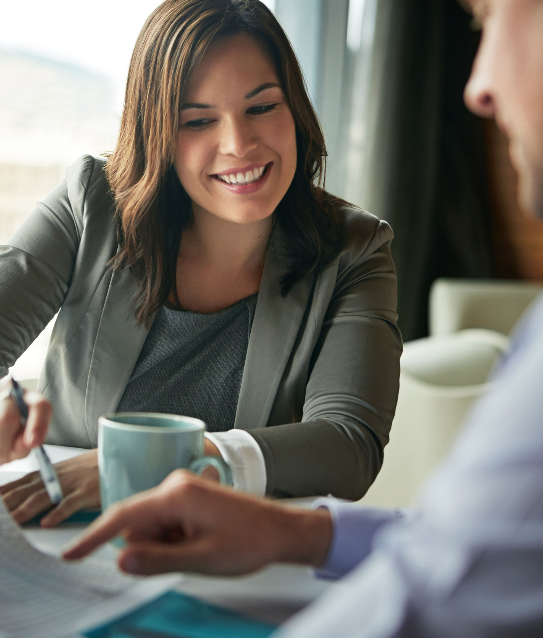 StockThingy1 A woman in business attire smiles while reviewing documents with a colleague at a table; a mug is beside her hand.